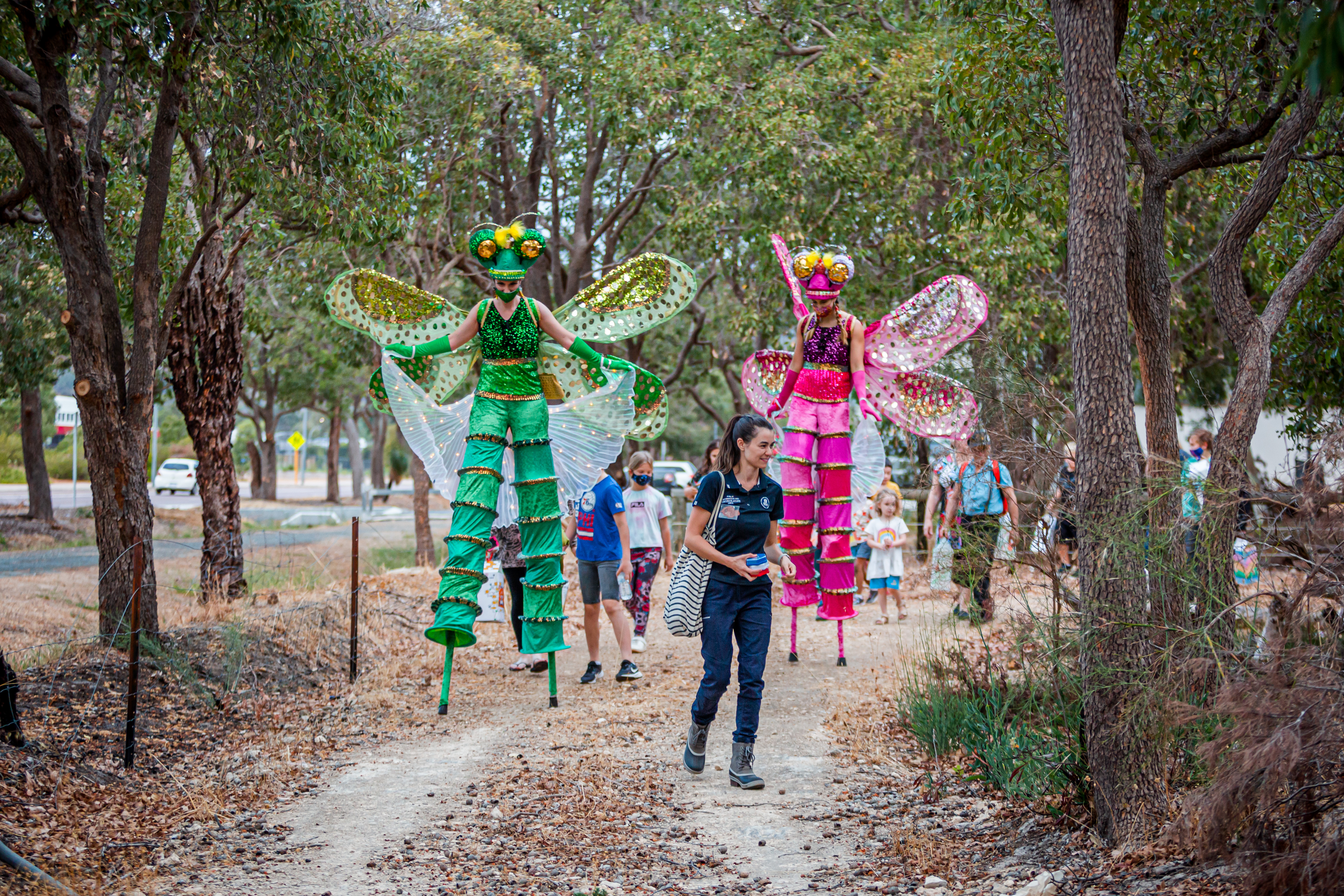 children and adults walk along a trail through the bush lead by two decorated stilt walkers