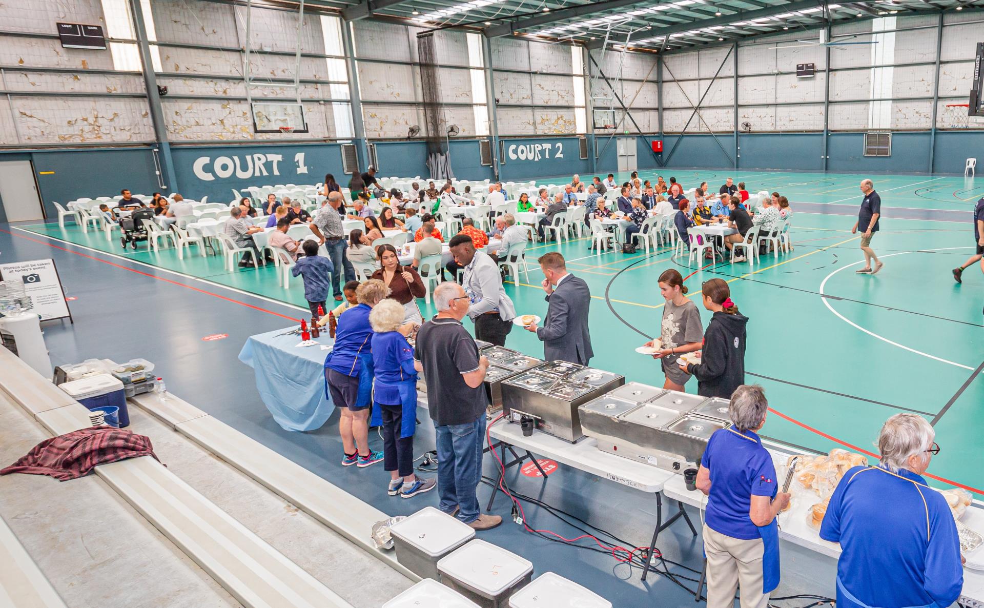 An indoor basketball court set with tables and chairs, people of all ages are seated at the tables. In the front right a buffet is set up and people are serving food.